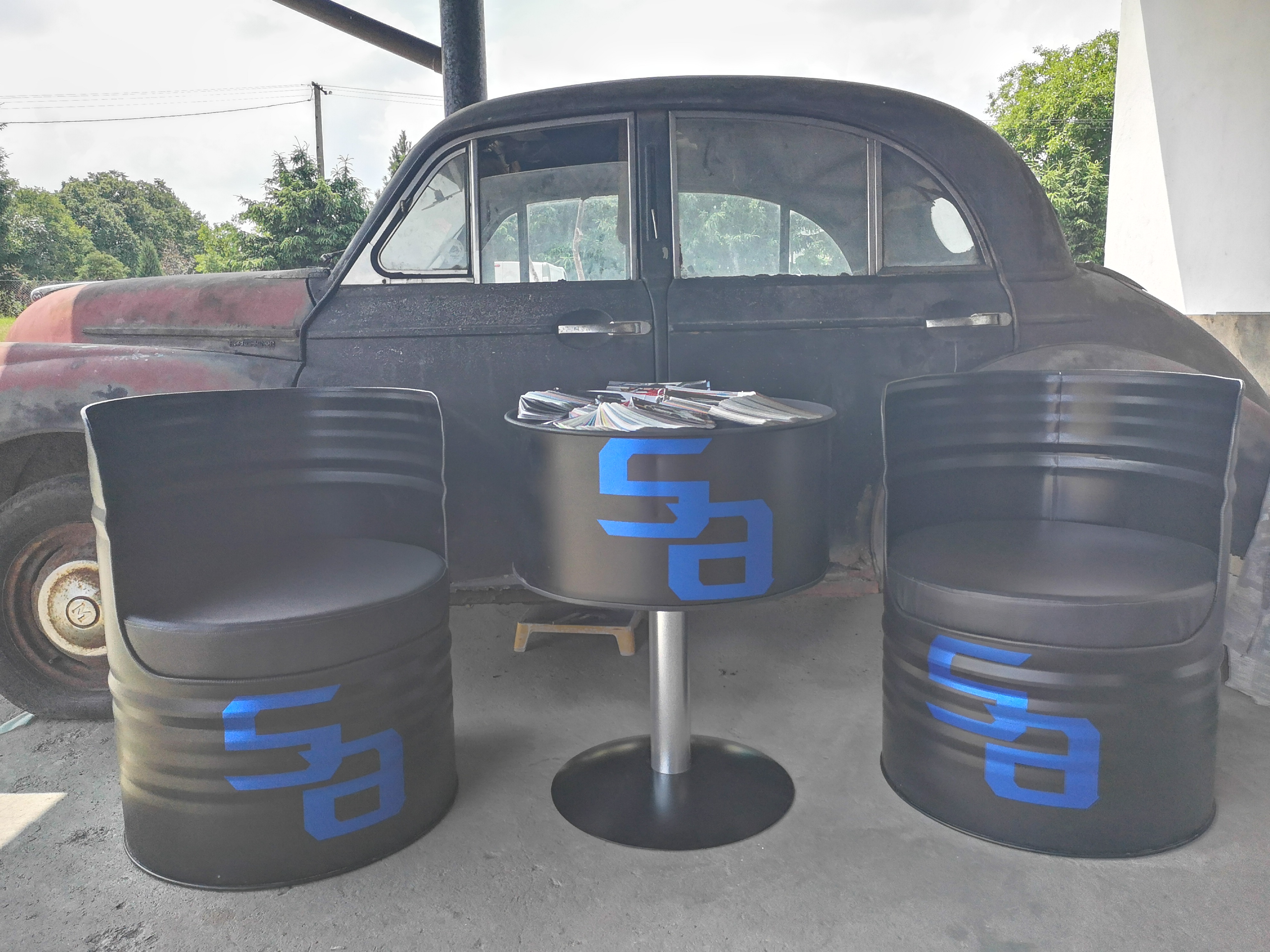 Używane auto marki Moskwicz w tle, a na pierwszym planie dwa krzesła i stolik wykonane z beczek z niebieskim logo.