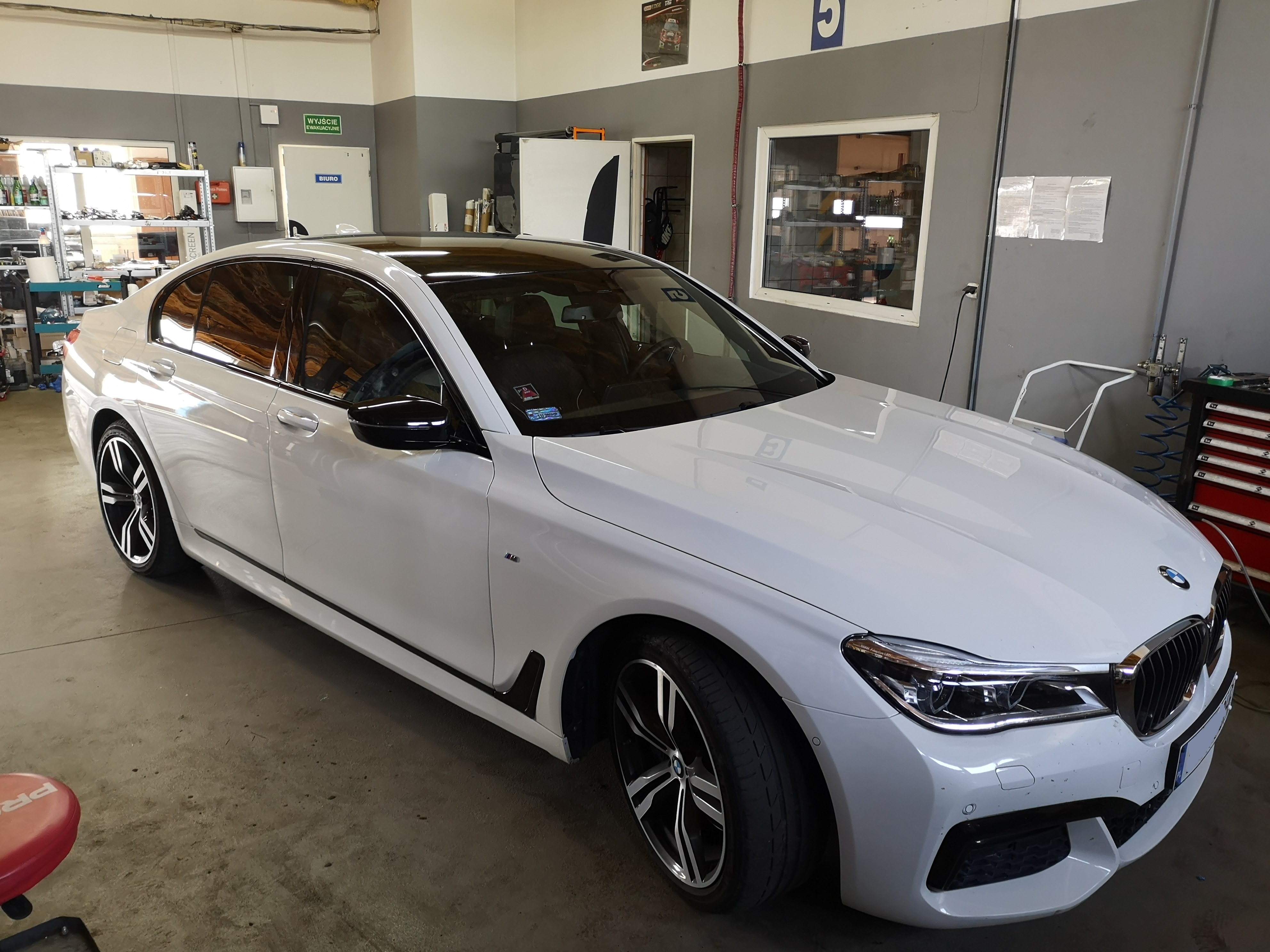 Biała limuzyna BMW serii 7 z czarnym dachem i przyciemnionymi szybami, zaparkowana w warsztacie samochodowym. Widoczne detale: czarne lusterka, felgi aluminiowe. W tle regały z narzędziami...