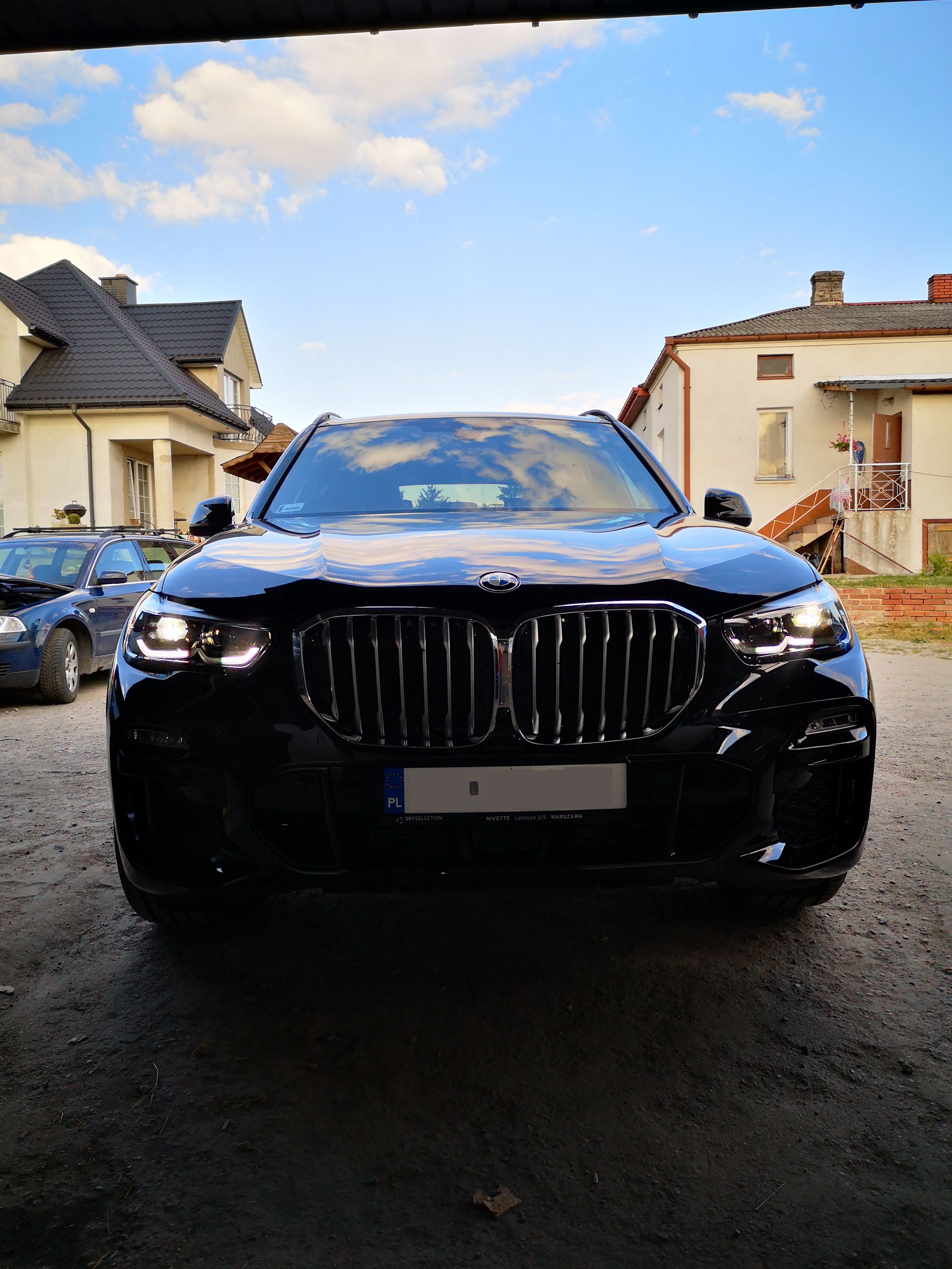 Czarny SUV BMW X5 z włączonymi światłami, zaparkowany na podwórku przed domem z odbiciem chmur na masce.