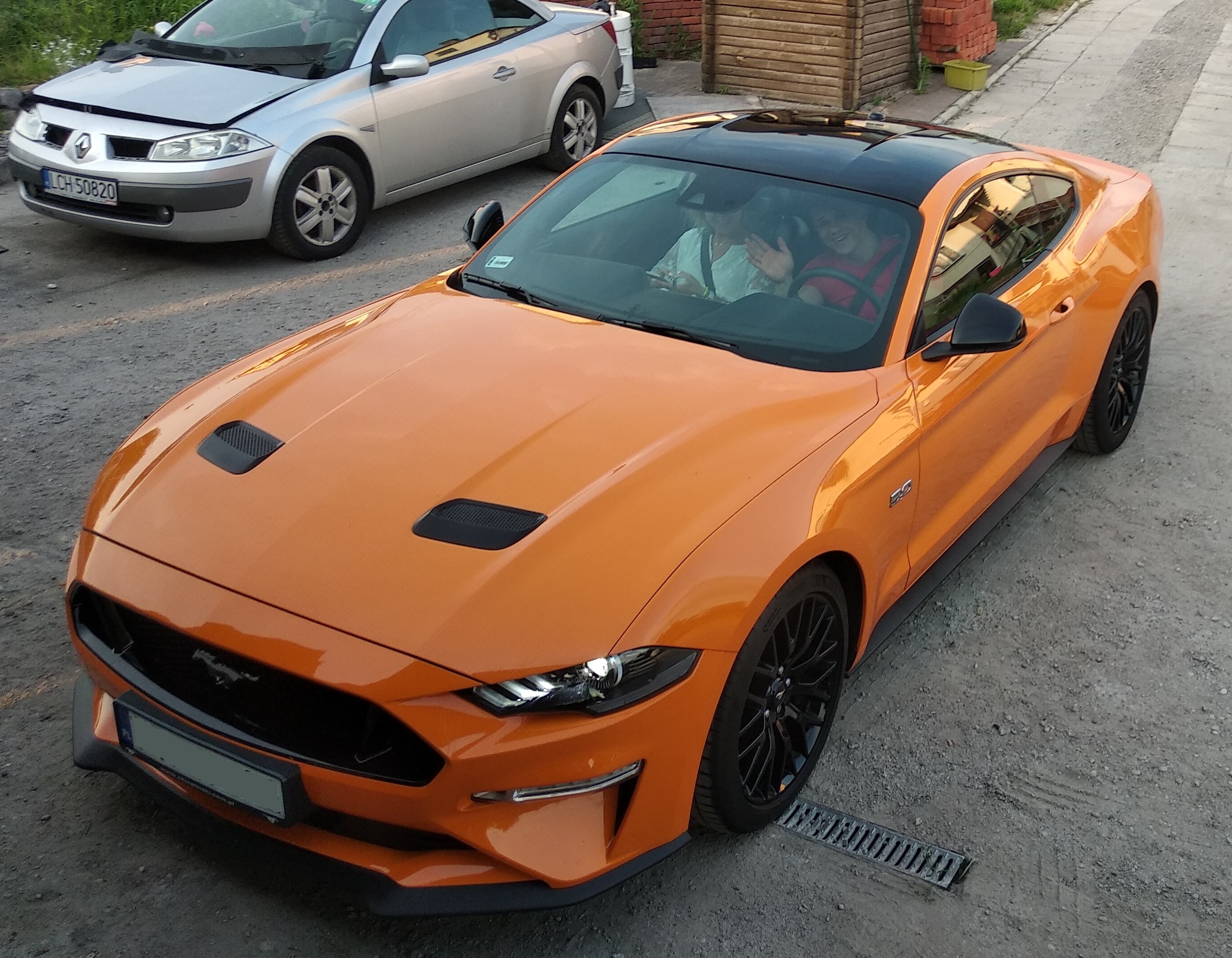 Pomarańczowy Ford Mustang z czarnym dachem, widziany z góry, z osobami w środku, zaparkowany obok srebrnego Renault Megane.