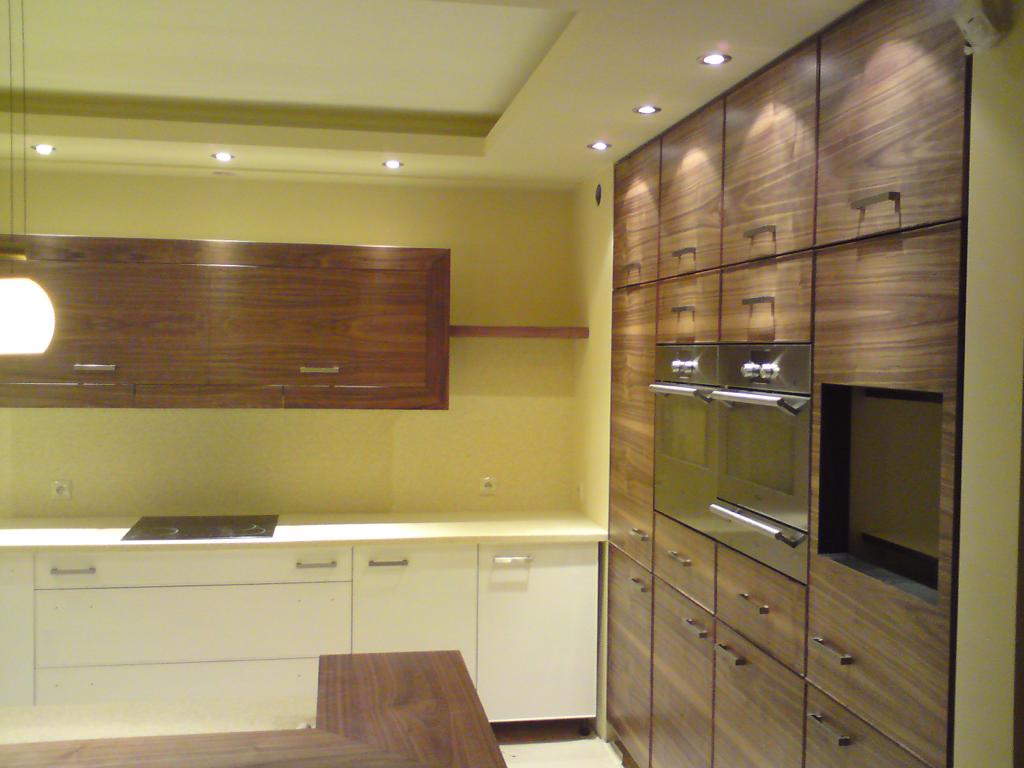 Nowoczesna kuchnia z drewnianymi frontami szafek, białymi blatami i wbudowanymi piekarnikami, oświetlona punktowymi lampami sufitowymi.