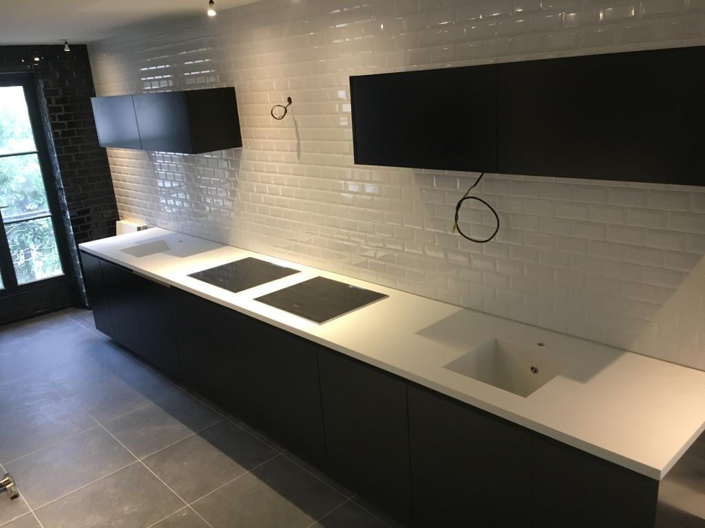 Nowoczesna kuchnia w trakcie wykończenia: czarne szafki dolne z białym blatem, białe płytki na ścianie, widoczne niedokończone podłączenie elektryczne, szara podłoga.