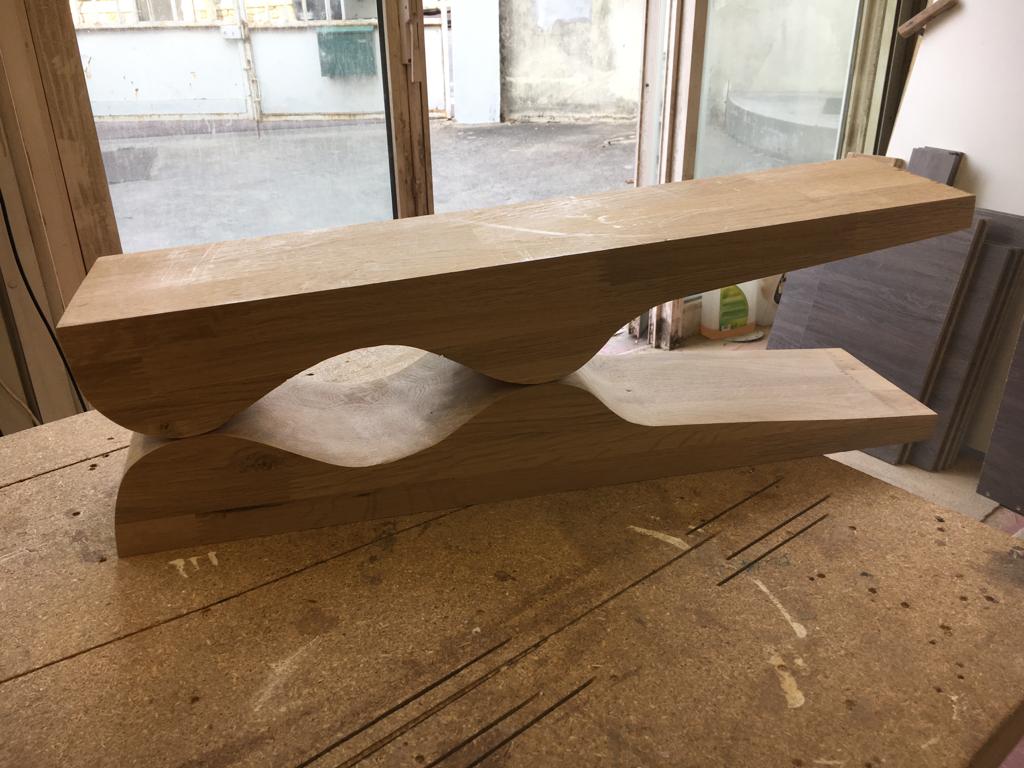 Surowy element meblowy z jasnego drewna, z wyciętymi falami, leżący na stole warsztatowym z widocznymi śladami obróbki, na tle okna i fragmentów płytek.