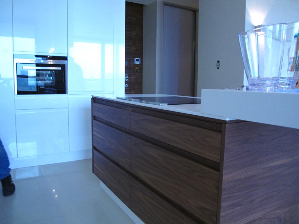 Nowoczesna kuchnia z białymi błyszczącymi szafkami i drewnianym blatem z szufladami, widoczny wbudowany piekarnik i płyta indukcyjna, minimalistyczny design.