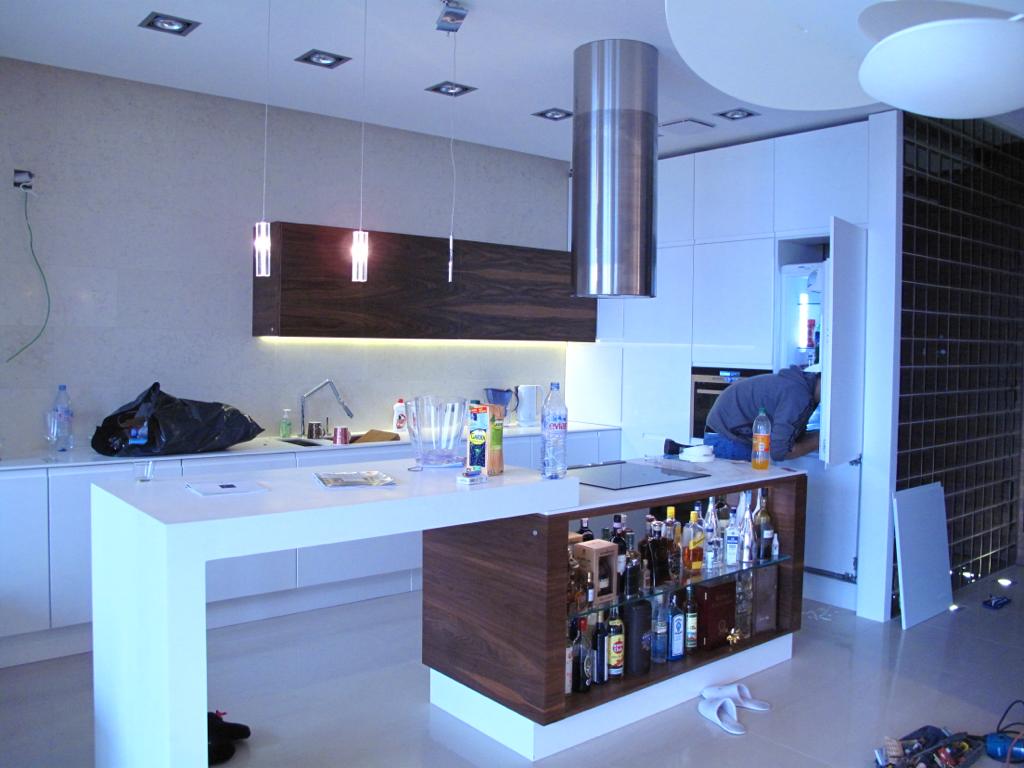 Nowoczesna kuchnia z białymi szafkami i wyspą z barkiem, z widocznym montażem lodówki przez osobę w szarej bluzie, z elementami wykończenia w drewnie orzechowym i minimalistycznym oświetleniem...