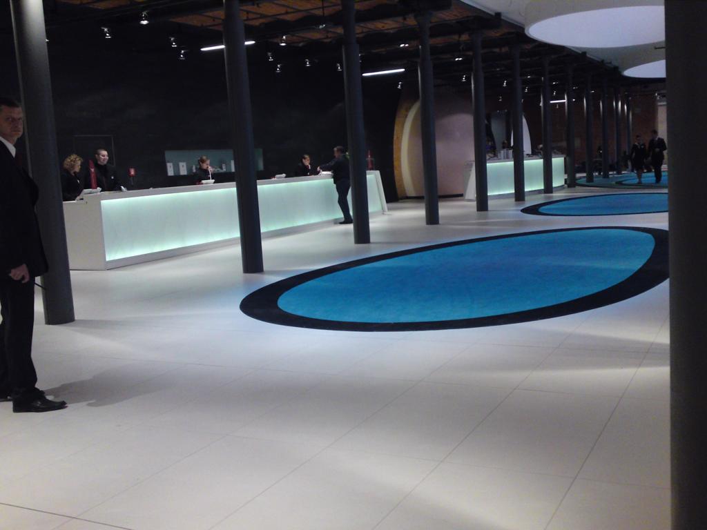 Nowoczesne wnętrze hali wystawienniczej z białymi podłogami, podświetlanymi ladami recepcyjnymi i okrągłymi, niebieskimi dywanami, widoczne ciemne kolumny podtrzymujące sufit.