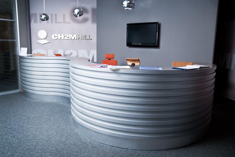 Recepcja biurowa z dwoma falistymi ladami w odcieniach szarości, logo firmy CH2MHILL na ścianie, nowoczesne lampy wiszące i telewizor LCD.