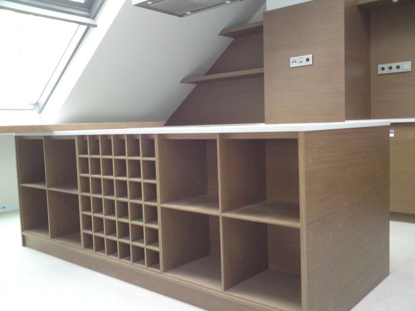 Brązowa zabudowa meblowa w skosie poddasza z otwartymi półkami i kwadratowymi przegródkami na butelki, widoczne gniazdka elektryczne na ścianie.