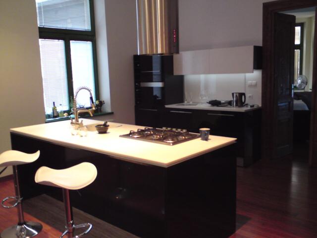 Nowoczesna kuchnia z wyspą: czarne szafki, biały blat, stalowy okap i dwa białe hokery. Widoczne okno z żaluzjami i drewniana podłoga.