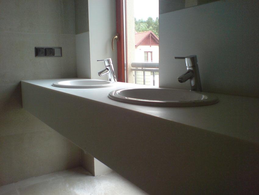 Dwie umywalki nablatowe z chromowanymi bateriami na blacie z jasnego konglomeratu w nowoczesnej łazience, widok przez okno na zewnątrz.