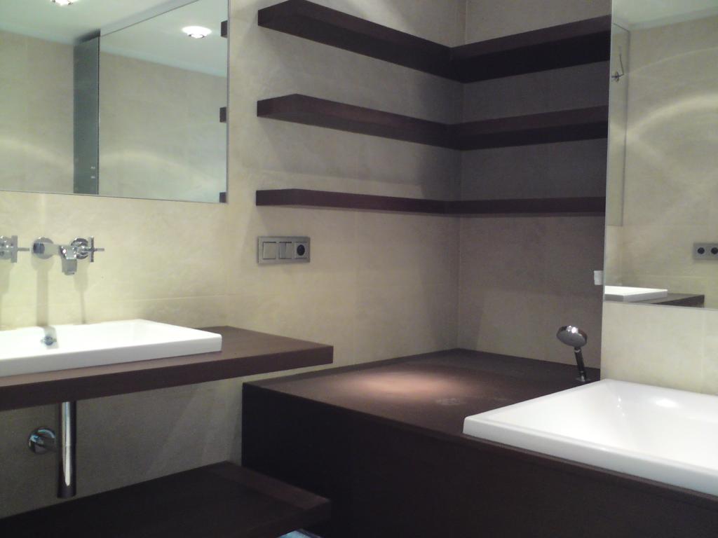 Nowoczesna łazienka z umywalką nablatową, wanną zabudowaną w ciemnym drewnie i półkami narożnymi w tym samym kolorze, podświetlana punktowo.