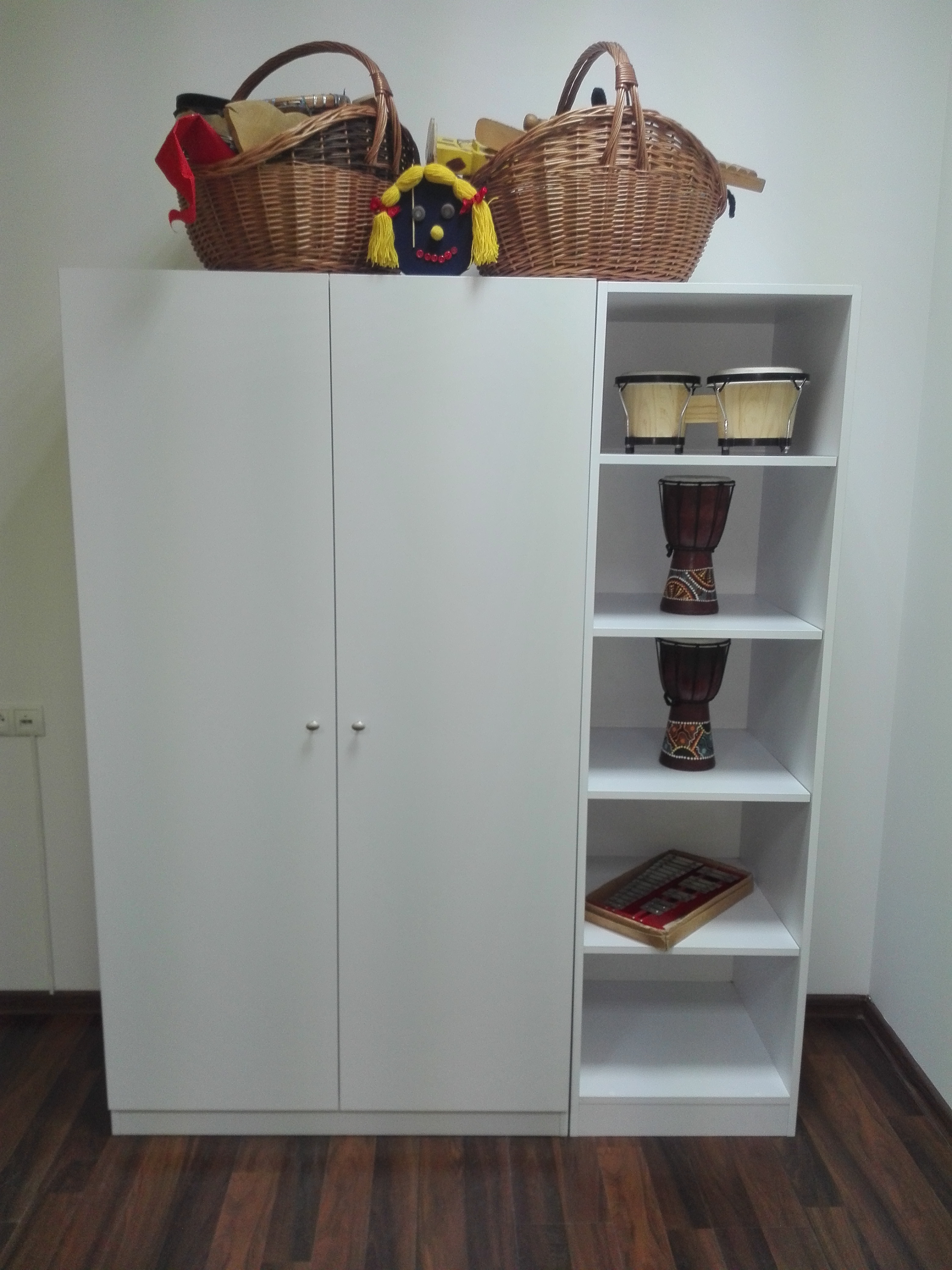 Biała szafa z drzwiami i otwartymi półkami, na których stoją bębny djembe i inne przedmioty, na górze szafy wiklinowe kosze i zabawka.