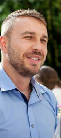 Portret uśmiechniętego mężczyzny w błękitnej koszuli, z delikatnym zarostem i srebrnym łańcuszkiem, spoglądającego w bok na tle zieleni.