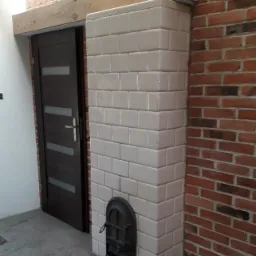 Kaflowy piec w trakcie budowy, z widocznymi białymi kaflami i żeliwnymi drzwiczkami, otoczony czerwoną cegłą i drewnianą belką stropową, z szufelką leżącą na podłodze.