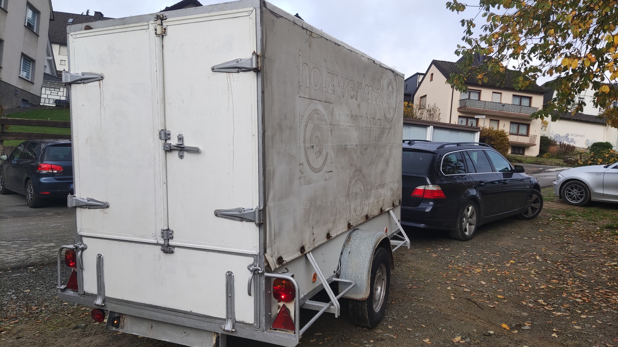 Biała przyczepa transportowa z plandeką, na której widnieje napis 'holzwerkstatt@' i grafika piły tarczowej, zaparkowana na żwirowym podłożu w otoczeniu samochodów i budynków.
