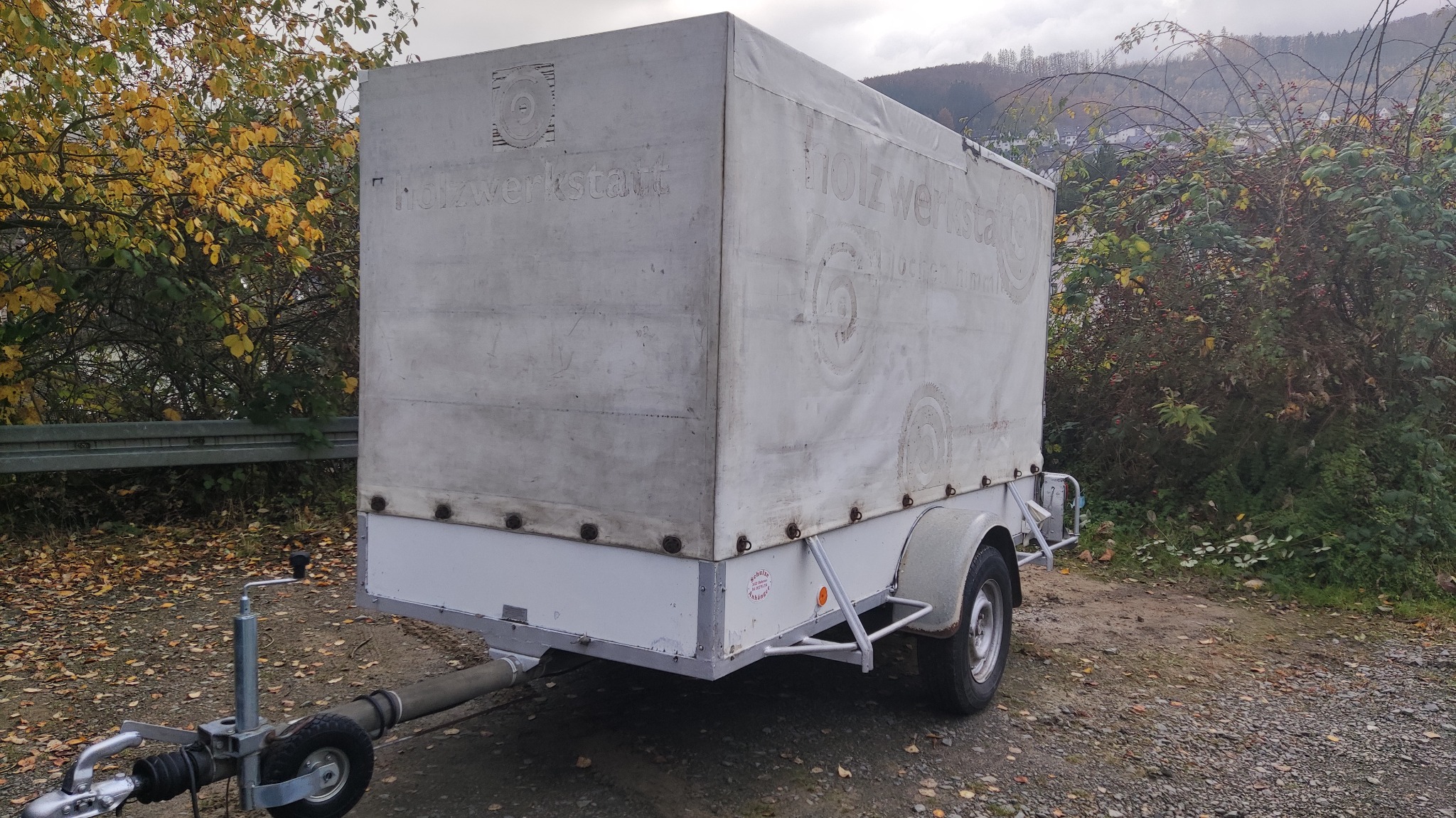 Przyczepa transportowa z plandeką, na której widnieje słabo widoczny napis 'Holzwerkstatt' oraz logo, zaparkowana na żwirowym podłożu, w tle drzewa z jesiennymi liśćmi.