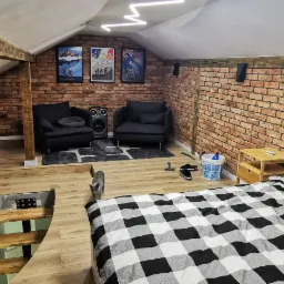 Zabudowa poddasza w Bielawie z widocznymi drewnianymi belkami, ceglaną ścianą, oświetleniem LED, dwoma fotelami i łóżkiem w kratę, ukazująca zaaranżowaną przestrzeń mieszkalną.
