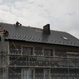 Ekipa dekarzy montuje ciemnoszarą dachówkę na domu jednorodzinnym, widoczne rusztowania i ocieplona elewacja budynku.