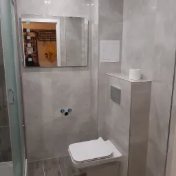 Nowoczesna łazienka w szarościach z podwieszaną toaletą, szklaną kabiną prysznicową i dużym lustrem na ścianie wyłożonej płytkami.