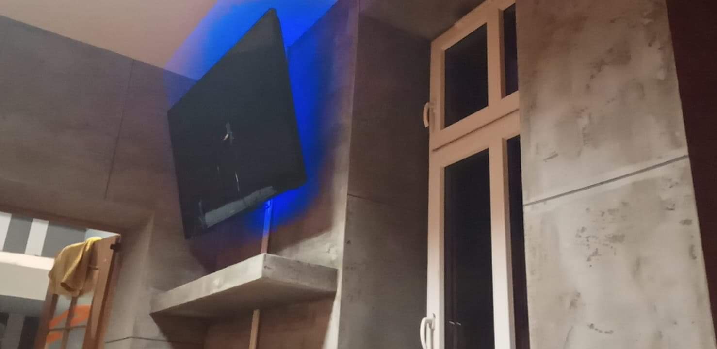 Surowe, betonowe ściany w salonie z oknem i telewizorem z podświetleniem LED, widoczne niedoskonałości i surowy charakter wykończenia.