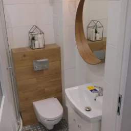 Nowoczesna łazienka z białą armaturą, umywalką na szafce, podwieszaną toaletą, okrągłym lustrem w drewnianej ramie i wzorzystą podłogą z płytek.