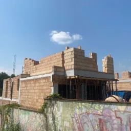 Wznoszenie ścian z bloczków ceramicznych na placu budowy, widoczne szalunki i zbrojenia, tło stanowi błękitne niebo z pojedynczą chmurą.