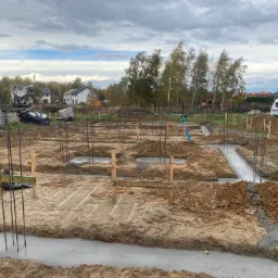 Wylane fundamenty pod budowę domu jednorodzinnego, widoczne zbrojenia, szalunki i betoniarki w tle, pochmurne niebo.