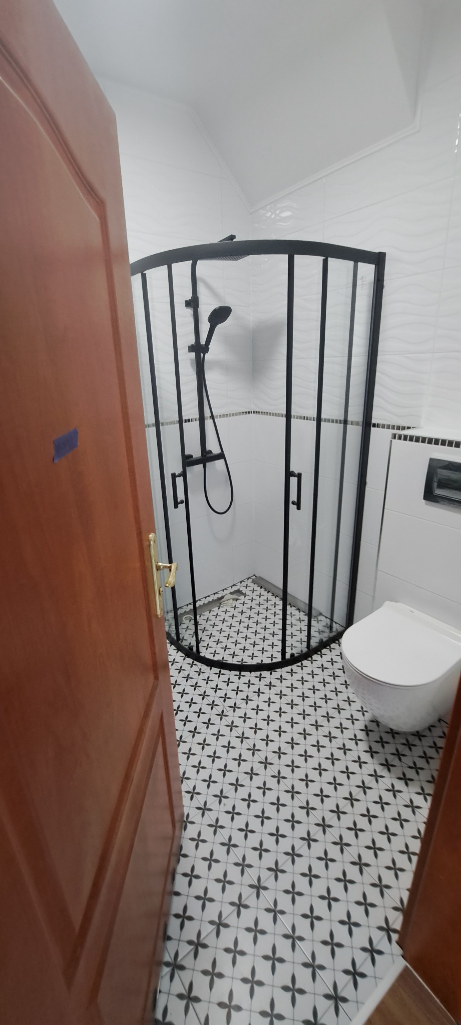 Wykończona łazienka z kabiną prysznicową narożną z czarnymi profilami, białą ceramiką i wzorzystą biało-czarną podłogą, widok zza uchylonych drewnianych drzwi.