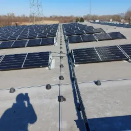 Rząd paneli fotowoltaicznych na płaskim dachu, widoczna instalacja elektryczna w korytach kablowych, maszty energetyczne w tle, cień fotografa w lewym dolnym rogu.