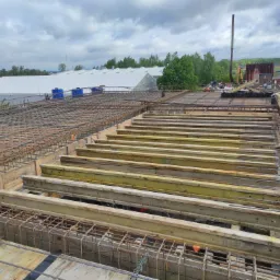 Zbrojenie stropu z drewnianymi belkami podtrzymującymi, widok z góry na konstrukcję z siatki stalowej na budowie, w tle budynki i drzewa pod pochmurnym niebem.