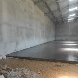 Wnętrze surowego budynku z betonową posadzką, oświetlone przez świetliki w metalowym dachu, z widocznym gruzem i piaskiem u podstawy ścian.