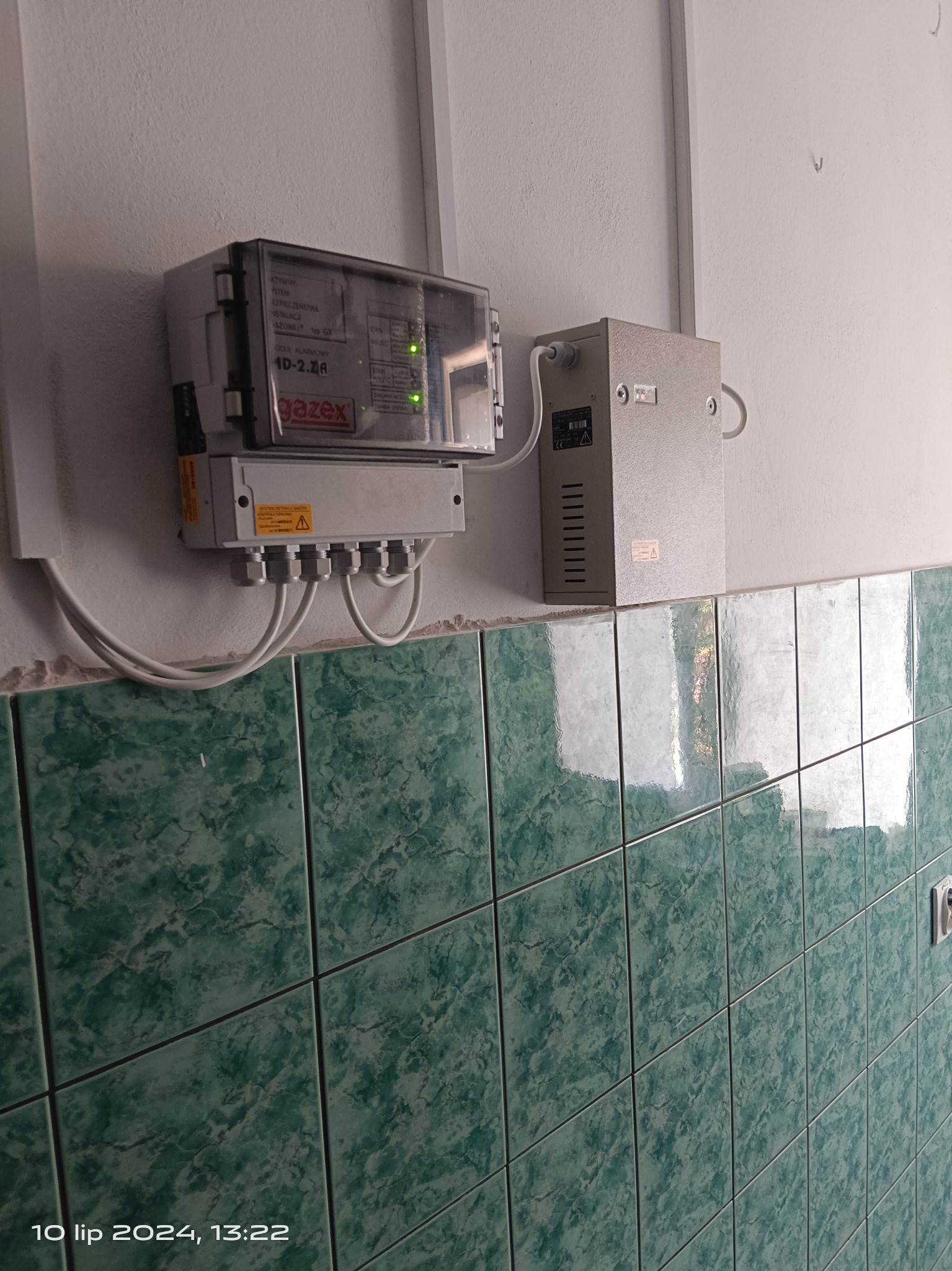 Zamontowana na ścianie centrala detekcji gazu Gazex z podłączonymi przewodami, obok znajduje się transformator. Widoczna zielona płytka ścienna z marmurkowym wzorem.