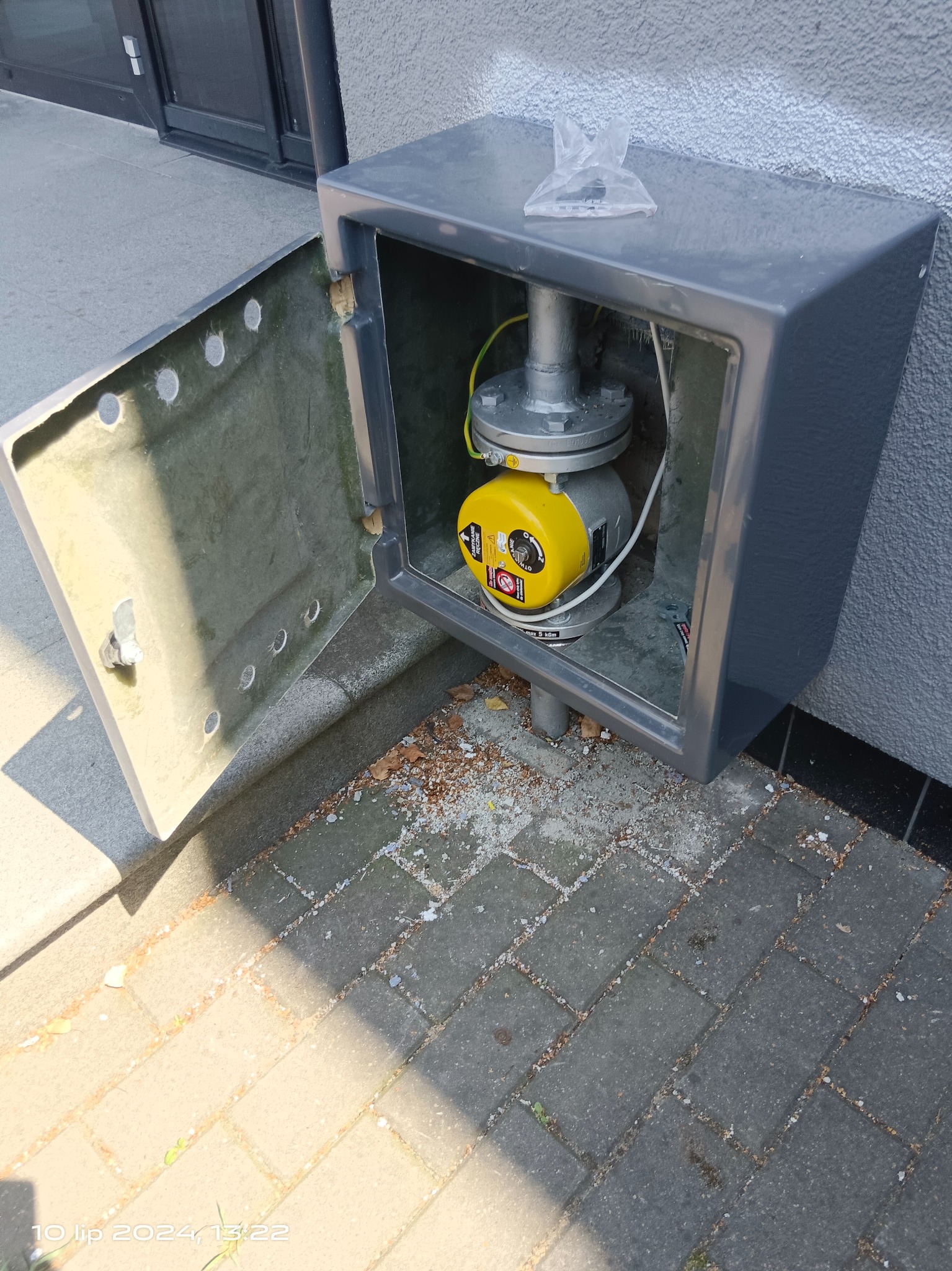 Zewnętrzna skrzynka gazowa z otwartymi drzwiczkami, widoczny żółty reduktor gazu, rury i złączki, na chodniku drobne zanieczyszczenia.