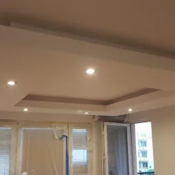 Biały, podwieszany sufit z wbudowanym oświetleniem punktowym, widok na okno z zasłoniętymi żaluzjami i wyjście na balkon.