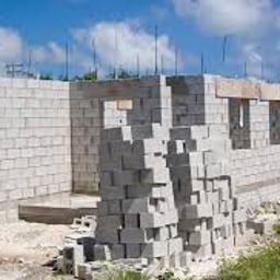 Kdkm - Wznoszenie ścian domu z bloczków betonowych, widoczne wystające pręty zbrojeniowe, stos bloczków przed budynkiem, błękitne niebo w tle.