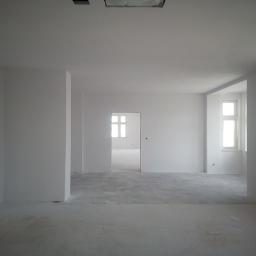 Kdkm - Wnętrze w stanie deweloperskim z surowymi ścianami pomalowanymi na biało, widoczne okna z białymi ramami, betonowa podłoga i otwór drzwiowy prowadzący do kolejnego pomieszczenia.