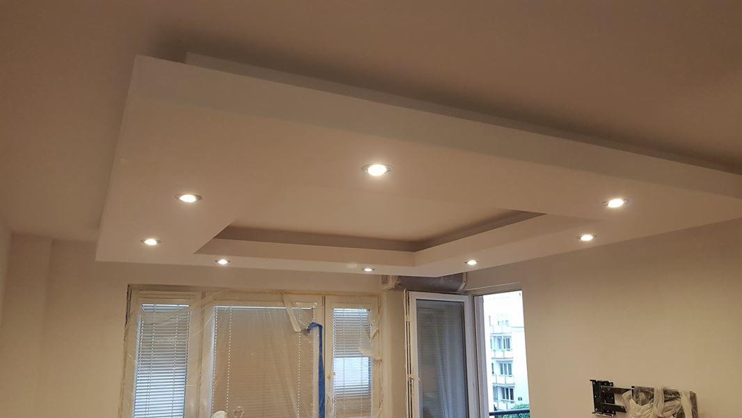 Biały, podwieszany sufit z wbudowanym oświetleniem punktowym, widok na okno z zasłoniętymi żaluzjami i wyjście na balkon.