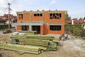 Budowa domu jednorodzinnego w stanie surowym otwartym: ściany z czerwonej cegły, widoczne drewniane elementy konstrukcji dachu, stos drewna budowlanego na pierwszym planie, w tle zabudowa...