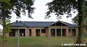 Niedokończony parterowy budynek mieszkalny z ciemnym dachem, widoczne surowe ściany z pustaków i filary, brak okien, teren porośnięty trawą, otoczony drzewami.