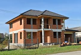 Dwukondygnacyjny dom w stanie surowym otwartym, zbudowany z czerwonej cegły, z widocznymi betonowymi słupami i przygotowanym zadaszeniem. Na pierwszym piętrze balustrada z metalowych prętów, w tle...