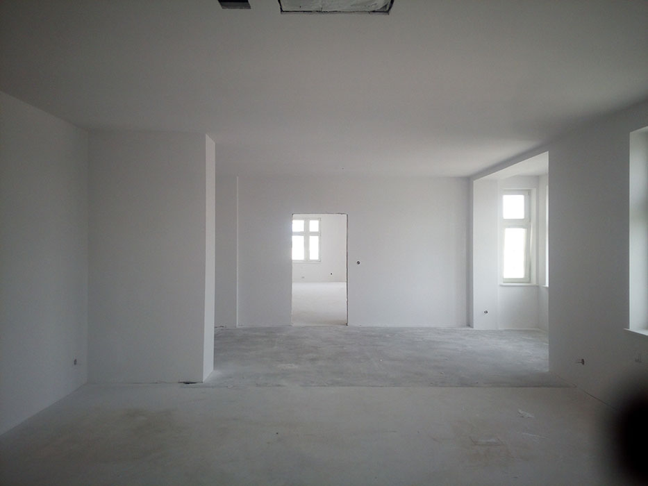 Wnętrze w stanie deweloperskim z surowymi ścianami pomalowanymi na biało, widoczne okna z białymi ramami, betonowa podłoga i otwór drzwiowy prowadzący do kolejnego pomieszczenia.
