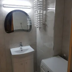 Nowoczesna łazienka z umywalką na szafce, grzejnikiem drabinkowym i pralką Bosch, oświetlona lampą nad lustrem o nietypowym kształcie.