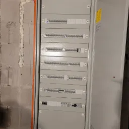 Otwarta szafa rozdzielcza z wieloma rzędami bezpieczników i wyłączników zamontowana na surowej ścianie z betonu komórkowego, uszczelniona pianką montażową.