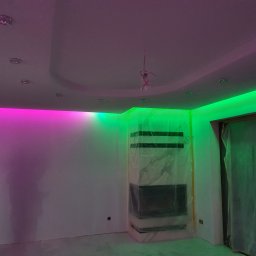 Usługi Elektryczne Piotr Borowski - Pokój w trakcie remontu z podświetleniem LED w kolorach różowym i zielonym, zabezpieczony kominek folią, osoba robiąca zdjęcie telefonem.
