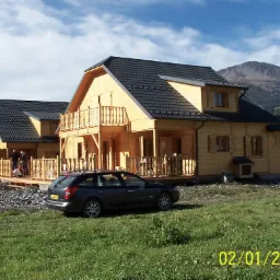 Dwa surowe, drewniane domy z ciemnymi dachami, jeden z balkonem, w otoczeniu trawy i gór, z samochodami zaparkowanymi w pobliżu.