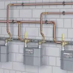 Instalacja gazowa: trzy liczniki gazu zamontowane na ścianie z białej cegły, połączone miedzianymi rurami z żółtymi zaworami.