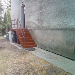 Zewnętrzne, pomalowane na brązowo schody z metalową barierką prowadzące do drzwi wejściowych budynku o szarej, chropowatej elewacji, widoczne ślady wilgoci na ścianie, obok stoją śmietniki.