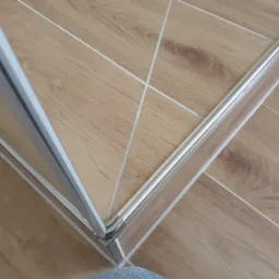 Szczegółowe ujęcie narożnika kabiny prysznicowej z aluminiowymi profilami na podłodze z paneli imitujących drewno z jasnymi fugami.