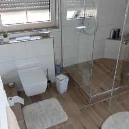 Nowoczesna łazienka z kabiną prysznicową typu walk-in, wiszącą toaletą i drewnopodobnymi płytkami podłogowymi, oświetlona naturalnym światłem z okna z żaluzjami.