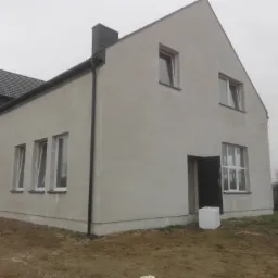 Surowy stan elewacji domu jednorodzinnego z widocznymi śladami tynkowania, otwartymi drzwiami wejściowymi i stertą styropianu obok, na tle pochmurnego nieba.